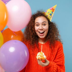 Photo of girl with a big smile and balloons, wearing a party hat and holding a cupcake.